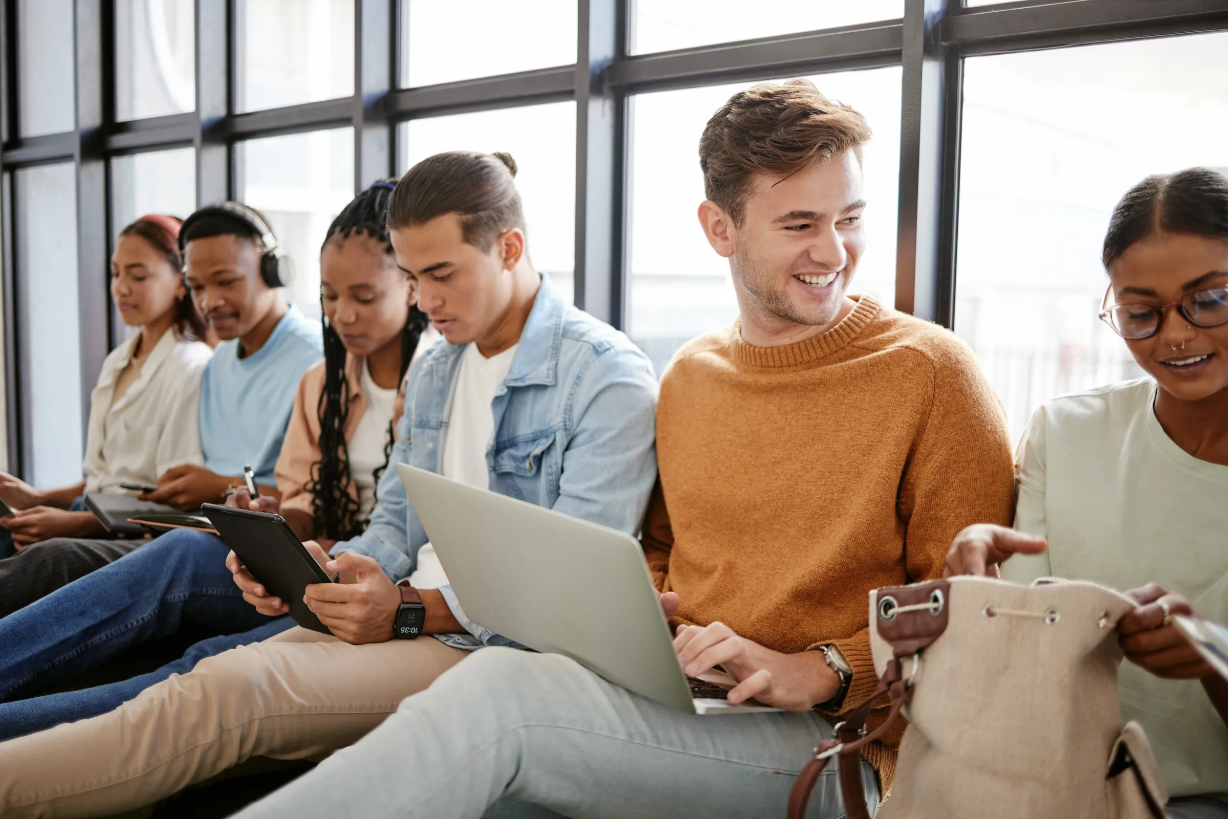 Studenten zitten naast elkaar bij een groot raam en werken op laptops en tablets